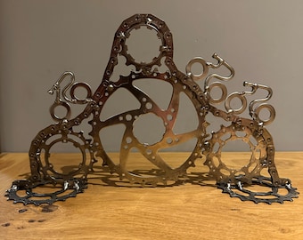 May include: A silver metal sculpture crafted from bicycle gears and chains, resembling a crown or decorative object. The artwork features various sizes and shapes of gears, creating an intricate and industrial design. The sculpture is displayed on a wooden surface.