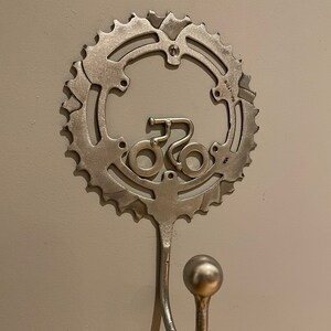 Puede incluir: Un gancho de pared de metal plateado con forma de rueda de bicicleta con una peque&ntilde;a silueta de bicicleta en el centro. El gancho est&aacute; fijado a una pared blanca.