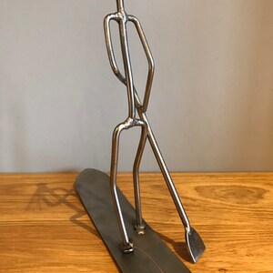 May include: A metal sculpture of a person standing on a surfboard with a paddle. The sculpture is made of silver metal and has a rustic, handmade look.