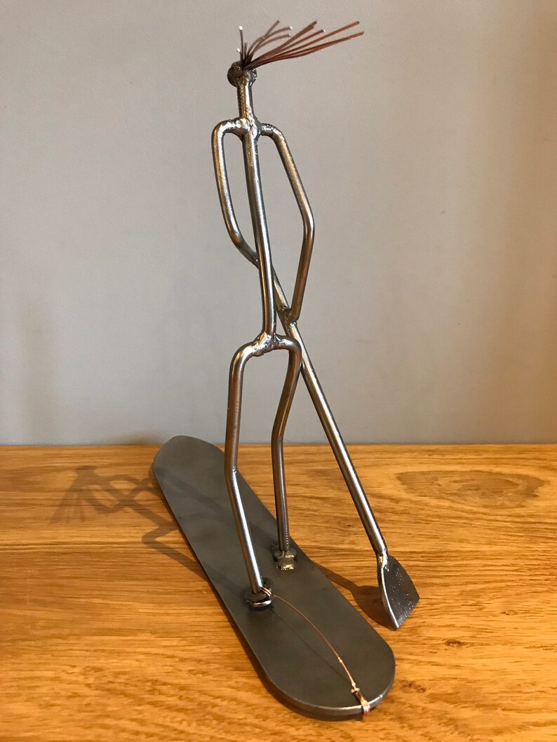 May include: A metal sculpture of a person standing on a surfboard with a paddle. The sculpture is made of silver metal and has a rustic, handmade look.