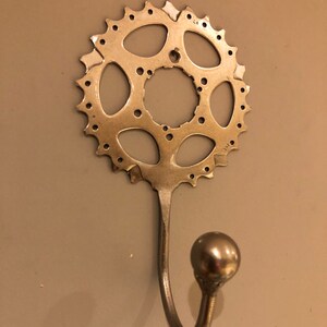 Puede incluir: Un gancho de metal plateado con una perilla redonda en el extremo. El gancho est&aacute; unido a un plato de cadena de bicicleta de metal plateado.