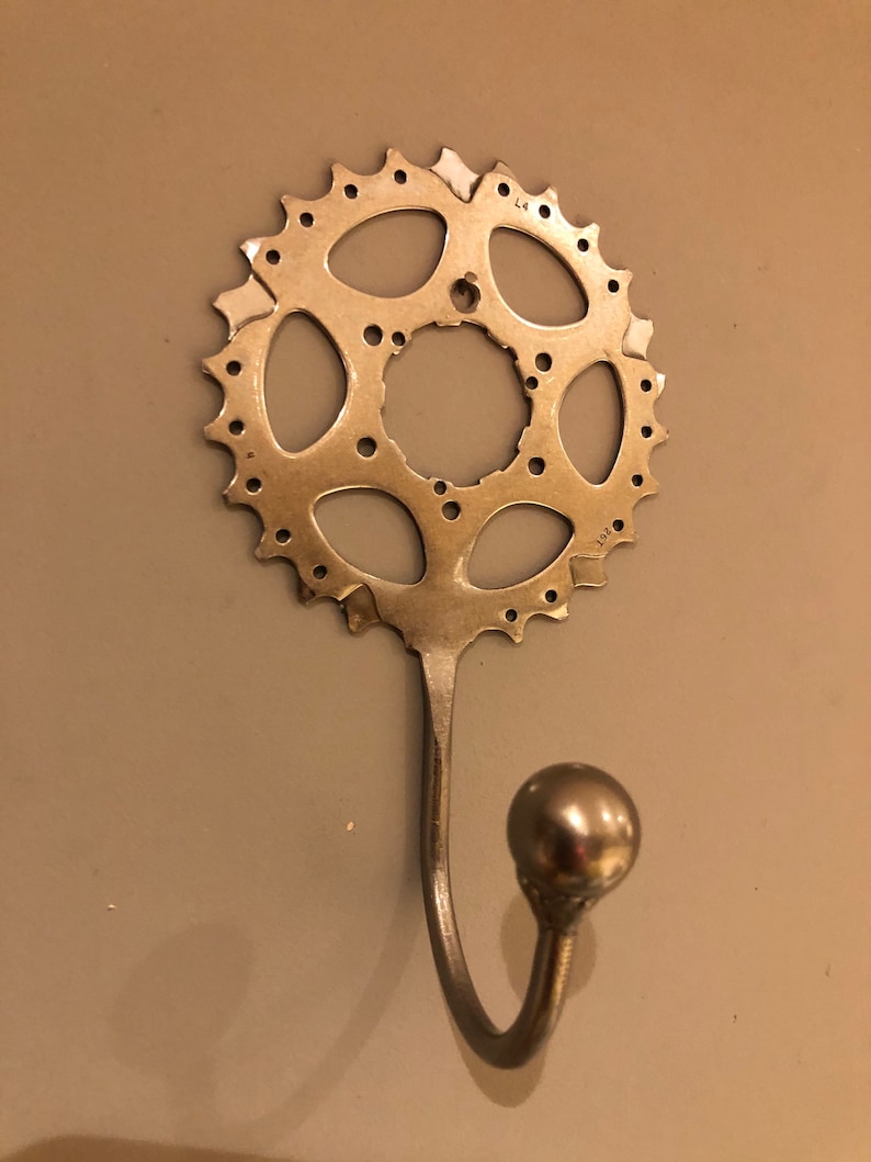 Puede incluir: Un gancho de metal plateado con una perilla redonda en el extremo. El gancho est&aacute; unido a un plato de cadena de bicicleta de metal plateado.