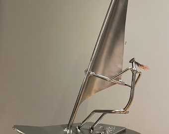 May include: A silver metal sculpture of a windsurfer. The sculpture is made of various metal pieces, including screws and bolts, and is mounted on a round base.