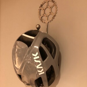 Puede incluir: Un casco de bicicleta gris con un logotipo KASK blanco colgado de un gancho de cadena de bicicleta de metal. El gancho est&aacute; fijado a una pared.