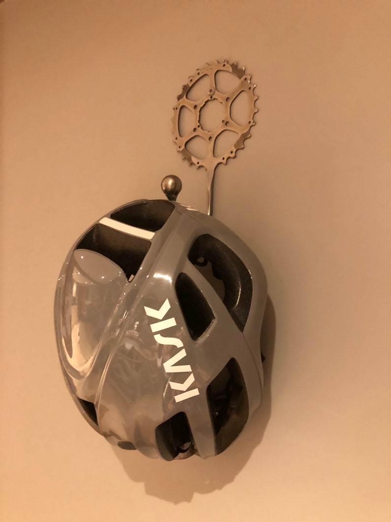 Puede incluir: Un casco de bicicleta gris con un logotipo KASK blanco colgado de un gancho de cadena de bicicleta de metal. El gancho est&aacute; fijado a una pared.