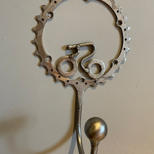 Puede incluir: Un gancho de pared de metal plateado con forma de bicicleta con un dise&ntilde;o de engranaje redondo.