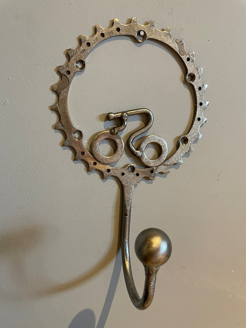 Puede incluir: Un gancho de pared de metal plateado con forma de bicicleta con un dise&ntilde;o de engranaje redondo.