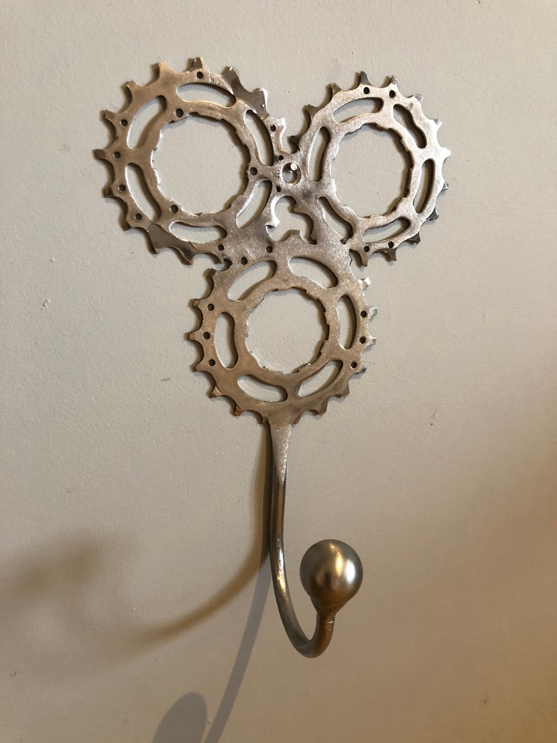 Puede incluir: Un gancho de pared de metal plateado con tres pi&ntilde;ones de cadena de bicicleta entrelazados. El gancho es curvo y tiene una perilla redonda en el extremo.