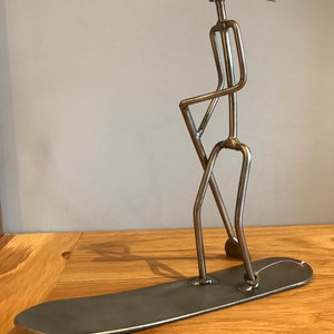 May include: A metal sculpture of a person standing on a surfboard. The sculpture is made of welded metal rods and has a copper wire hairpiece. The sculpture is a unique and whimsical piece of art.
