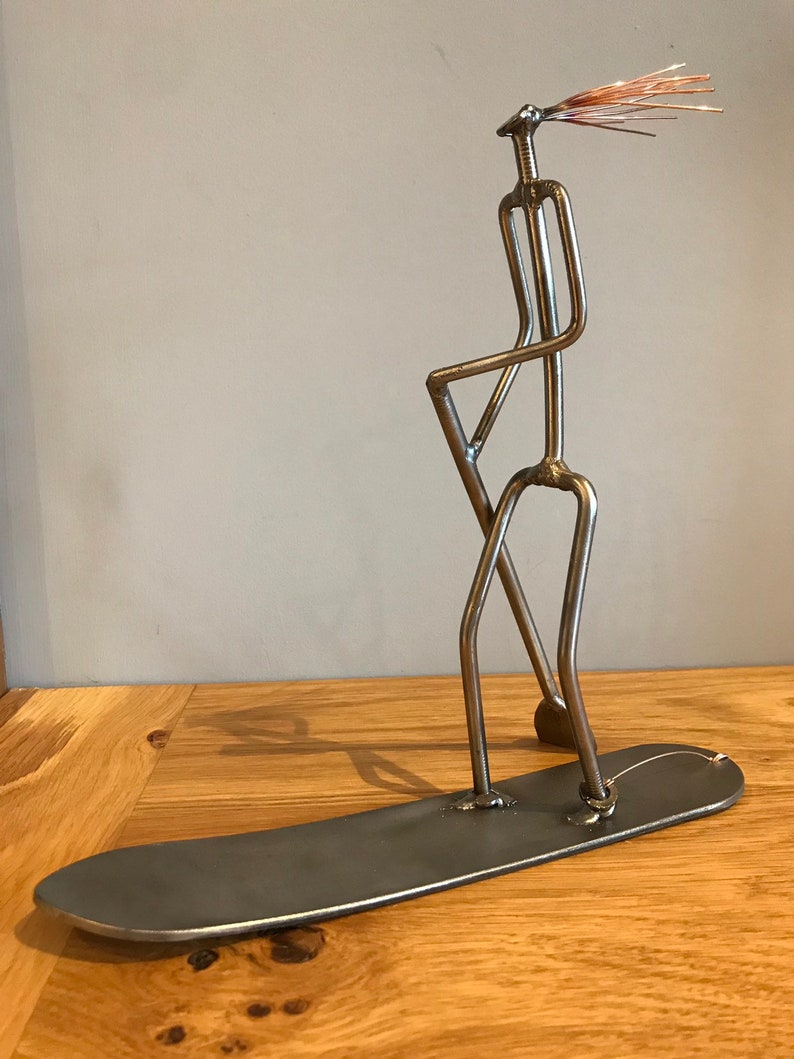 May include: A metal sculpture of a person standing on a surfboard. The sculpture is made of welded metal rods and has a copper wire hairpiece. The sculpture is a unique and whimsical piece of art.