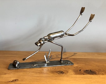 May include: A handcrafted metal sculpture of a diver, made from repurposed metal. The diver is bent over, with arms outstretched, and is attached to a rectangular base. The sculpture is silver in color and sits on a wooden surface.