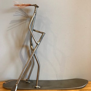 May include: A metal sculpture of a person standing on a surfboard, holding a paddle. The sculpture is made from recycled metal and has a rustic, industrial look.