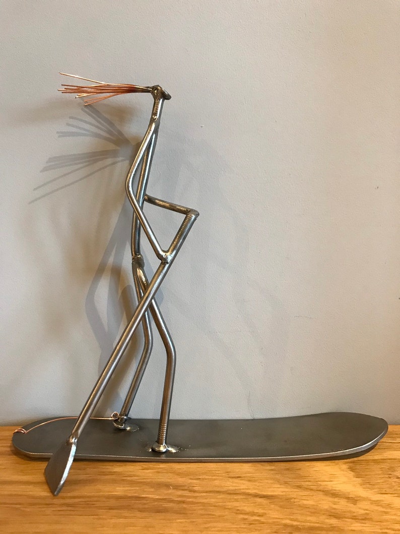 May include: A metal sculpture of a person standing on a surfboard, holding a paddle. The sculpture is made from recycled metal and has a rustic, industrial look.