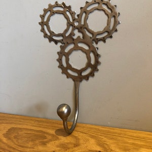 Puede incluir: Un gancho de pared de metal plateado con tres engranajes entrelazados. El gancho est&aacute; unido a la parte inferior de los engranajes.