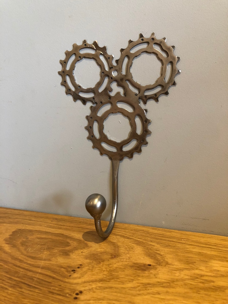 Puede incluir: Un gancho de pared de metal plateado con tres engranajes entrelazados. El gancho est&aacute; unido a la parte inferior de los engranajes.