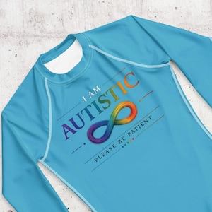 Op de afbeelding: Een lichtblauw shirt met lange mouwen en een regenboogkleurig oneindigheidssymbool. De tekst luidt I AM AUTISTIC en PLEASE BE PATIENT in donkerblauwe letters. Het ontwerp bevat horizontale lijnen en een reeks kleine gekleurde stippen.