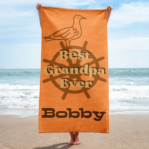 Puede incluir: Toalla de playa naranja con una gaviota marrón y un timón marrón con el texto "Best Grandpa Ever" y el nombre "Bobby" en texto marrón.