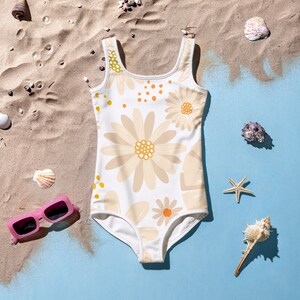 May include: White one-piece swimsuit with a floral pattern in beige and yellow. The swimsuit features a scoop neck and a low back. The image is set on a beach with seashells and pink sunglasses.