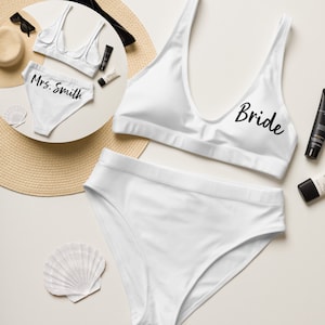 Puede incluir: Conjunto de bikini blanco con las palabras "Bride" y "Mrs. Smith" impresas en las partes de arriba. El bikini está colocado sobre una superficie blanca con un sombrero de paja y conchas.