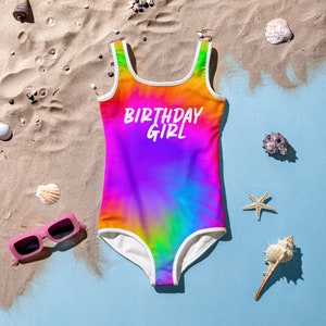 Puede incluir: Un traje de ba&ntilde;o de te&ntilde;ido anudado arco&iacute;ris con ribetes blancos y el texto "Birthday Girl" en la parte delantera. El traje de ba&ntilde;o est&aacute; sobre una playa de arena.