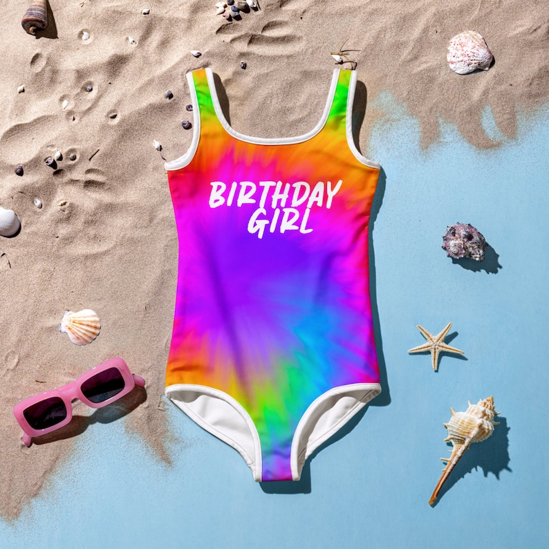 Puede incluir: Un traje de ba&ntilde;o de te&ntilde;ido anudado arco&iacute;ris con ribetes blancos y el texto "Birthday Girl" en la parte delantera. El traje de ba&ntilde;o est&aacute; sobre una playa de arena.