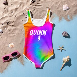 Puede incluir: Un traje de ba&ntilde;o de tie-dye arco&iacute;ris con ribetes blancos y el nombre "QUINN 3" impreso en blanco en la parte delantera.