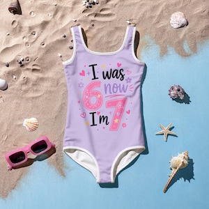 May include: A light purple one-piece swimsuit with white trim featuring the text I was 6 now I'm 7 in black and pink lettering. The design includes small pink and yellow hearts, stars, and flower graphics scattered around the central numbers.