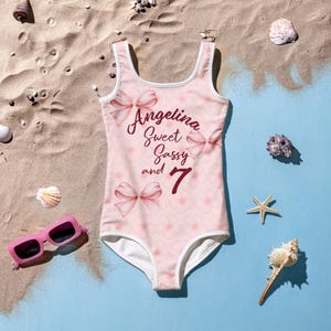 Può includere: Costume da bagno rosa con bordino bianco e motivo floreale. Il costume da bagno ha la scritta "Angelina Sweet Sassy and 7" stampata.