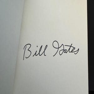 May include: A page from a book with the handwritten signature of "Bill Gates" in black ink. The signature is centered on the white page, with the book open to reveal the signed page.