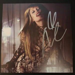 May include: A photograph featuring a person with long, light brown hair and dark eye makeup. The person is wearing a black, net-like garment. The image has a signature in white. The background is blurred with warm tones.