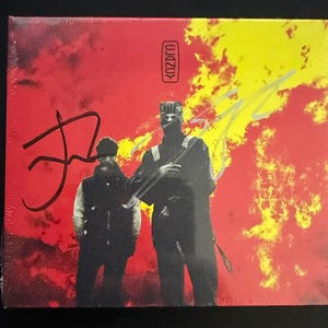 May include: A square music album cover featuring a red and yellow background with two silhouetted figures in dark clothing. The text KOZDRO is printed in a vertical box at the top center. Two distinct signatures are written in black and silver ink.
