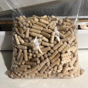 May include: A clear plastic bag filled with a large quantity of small, wooden dowels. The dowels are all the same size and are arranged in a random pattern. The dowels are a light brown color.
