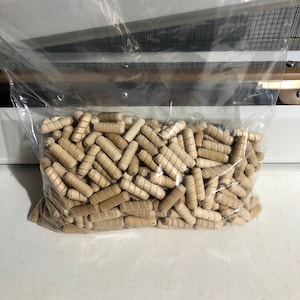May include: A clear plastic bag filled with a large quantity of small, wooden dowels. The dowels are light brown in color and have a slightly textured surface.