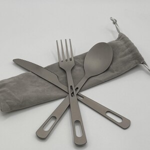 May include: A gray, four-piece travel utensil set including a knife, fork, spoon, and a carrying pouch. The utensils are made of a matte metal and have a minimalist design with a hole in the handle. The pouch is a soft gray fabric.