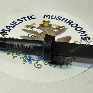 May include: A black drill bit with a 8.5 mm diameter, featuring a screw tip and a hex shank. The drill bit is resting on a white sticker with a mushroom design and the text "Majestic Mushrooms".