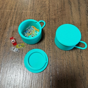 May include: A teal-colored sewing clip storage container with a lid and handle. The container is open, revealing several colorful sewing clips. The lid is off to the side. The container is on a wooden surface.