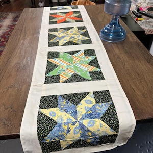 May include: A rectangular table runner with four star-shaped quilt blocks. The stars feature various patterns and colors, including orange, yellow, green, and blue, set against a black background. The runner is bordered with a cream-colored fabric.