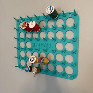 May include: A teal thread spool holder with multiple pegs and circular openings, holding various spools of thread in red, green, and yellow. The words "SEW MORE" are printed on the holder.