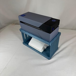 May include: A rectangular thermal label printer with a gray and black casing sits atop a blue 3D-printed plastic stand. The stand features vertical cutouts on its sides and holds a stack of white folded labels underneath the printer unit.