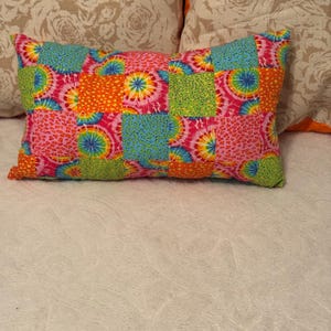 May include: A rectangular patchwork pillow with a vibrant mix of colors. The pillow features squares of pink, orange, blue, and green fabric with tie-dye and leopard print patterns. The pillow is resting on a white surface.