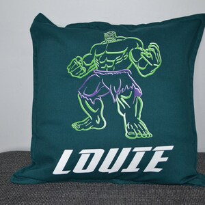 May include: Teal cushion featuring a green and purple embroidered image of the Hulk. The word "LOUIE" is embroidered in white block letters across the bottom. The cushion is square and rests on a grey surface.
