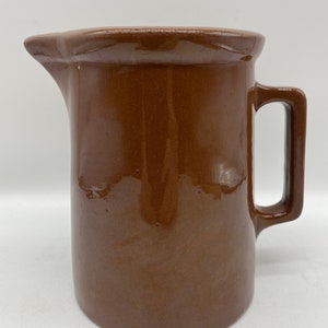 Puede incluir: Una jarra de cerámica marrón con asa. La jarra tiene un acabado liso y brillante.