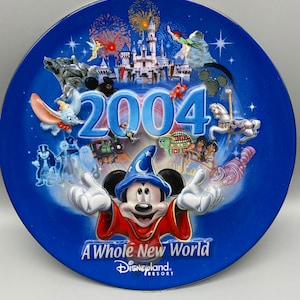 May include: A blue ceramic plate featuring a colourful illustration of Mickey Mouse as a sorcerer, surrounded by images of Disneyland attractions. The plate displays the year 2004 and the text "A Whole New World" and "Disneyland Resort".