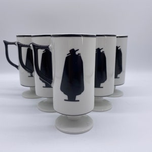 May include: Set of six white ceramic mugs with black trim and a black silhouette of a person wearing a hat and a cape. The mugs have a pedestal base.