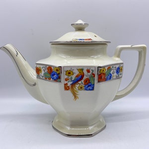 May include: A white teapot with a floral and bird design. The teapot has a lid with a silver trim and a silver trim around the base. The teapot has a octagonal shape.