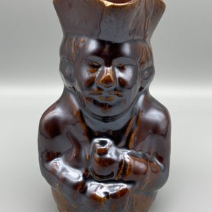 Puede incluir: Un jarro de cerámica marrón con forma de persona que lleva un sombrero. La persona sostiene una pipa en la mano.