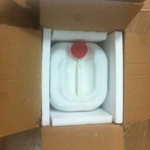 Könnte beinhalten: Ein weißer Plastikbehälter mit rotem Deckel ist in einem Karton mit Styropor-Polsterung verpackt.