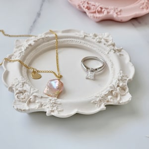 Puede incluir: Un plato de cer&aacute;mica blanca con un dise&ntilde;o ornamentado, que presenta un collar de oro con un colgante de perla rosa y un anillo de plata con un diamante transparente.