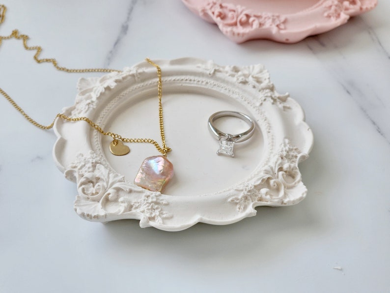Puede incluir: Un plato de cer&aacute;mica blanca con un dise&ntilde;o ornamentado, que presenta un collar de oro con un colgante de perla rosa y un anillo de plata con un diamante transparente.