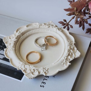 Puede incluir: Plato de cer&aacute;mica blanca para anillos con detalles ornamentados, que presenta tres anillos: una banda dorada, una banda plateada y una banda plateada con un diamante de talla cuadrada.